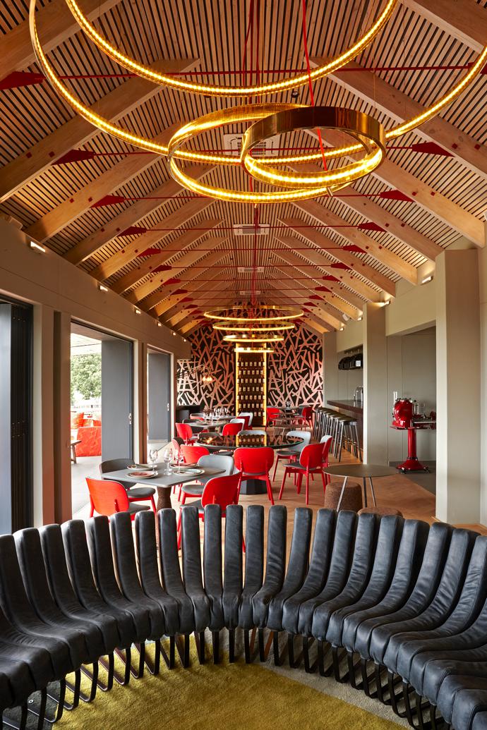 Brass halo lights lead the eye to the Sarah Ord Crayolla wallpaper, its bold brushstroke pattern echoing the Kunjani! Wines logo. Haldane Martin Sim-ply chairs add pops of accent red.