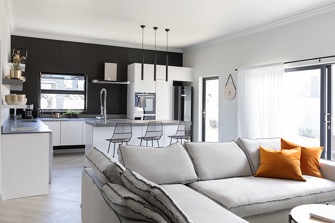 The open-plan lounge and kitchen area was planned and executed to perfection. The soft grey L-shaped sofa from Weylandts is in harmony with the predominant Plascon Nomadic Dream (EC56) paint shade and Belgotex Charleston wood-look vinyl flooring used throughout the house. In the background, Plascon School Board Paint, Caesarstone tops, Grohe taps, Grundig appliances and black tube pendant lights from Spazio Lighting contributed to this space winning the kitchen challenge. The wire barstools are from Red Apple Furniture, the clock is from @home, the Kambrook small appliances are from Clicks and the rest of the accessories are from H&M Home.