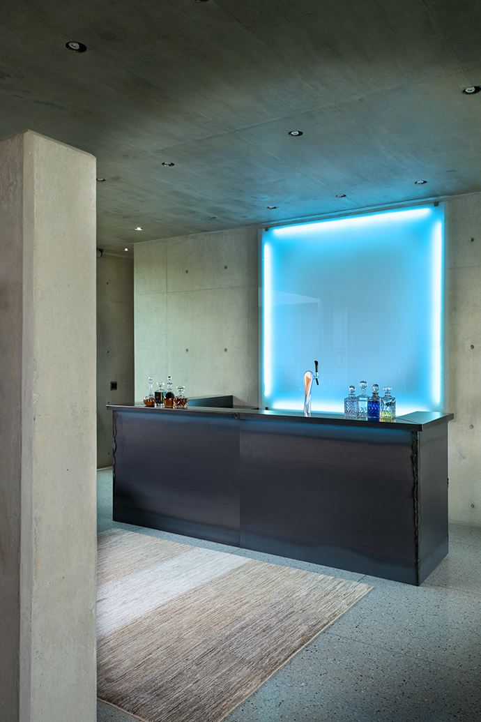 The bar is clad in mild steel, which was oiled and then fired to give it a distressed look. Behind the bar, a sheet of white glass backlit with a blue light emits a cool glow.