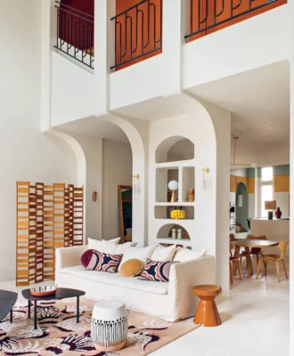 Benefiting from a triple volume, and animated by vertical and horizontal flows, the living room forms the centrepiece of this apartment’s design. It offers views to the other spaces of the triplex, with structural elements softened by plaster curves and built-in joinery in organic shapes. A balcony overlooks this vast shared room, and the kitchen can be seen in the background.