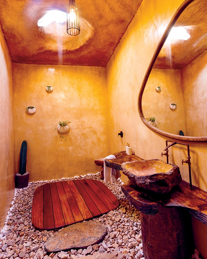 Below a Meyer Von Wielligh mirror, a stone basin carved from local rock sits on a timber slab, also by Meyer Von Wielligh, made from the wood of a fallen English oak. Shower water falls from the open skylight.