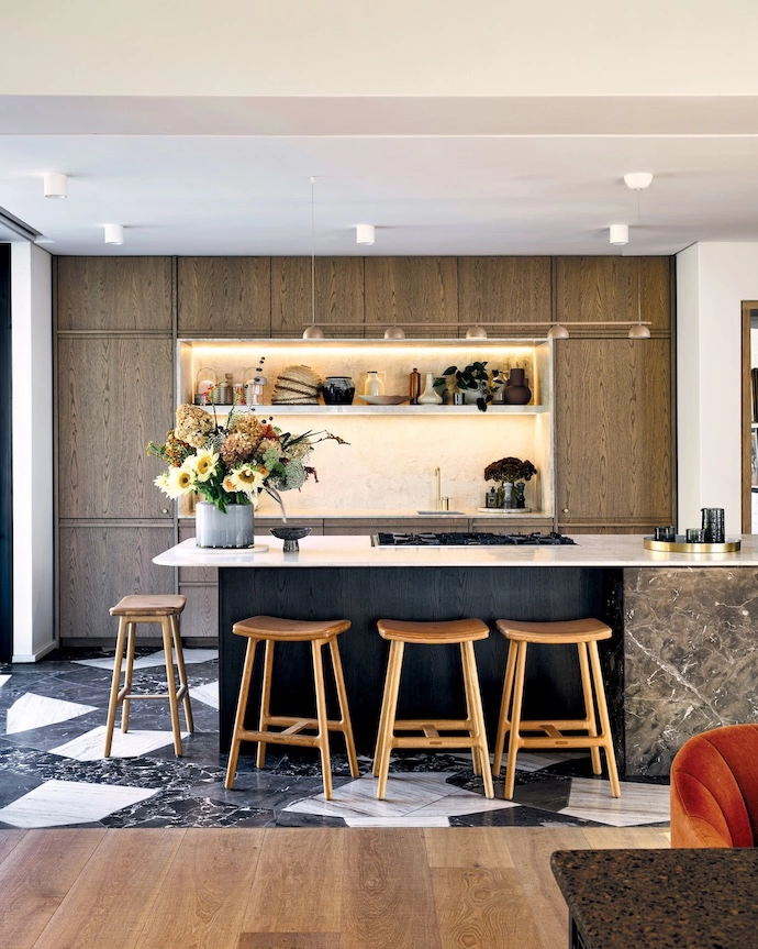 In the kitchen, a geometric marble floor by Continua connects with the marble countertop from Mazista Tiles and the marble inlaid shelving on the back wall. The mix is sophisticated against the wooden cabinetry. The barstools are from Weylandts.