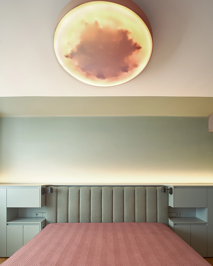 The bedroom continues the calm palette of the apartment — pink, green, and natural wood create a balanced space for rest. A custom ceiling light evokes the glow of a setting sun, softening the atmosphere.
