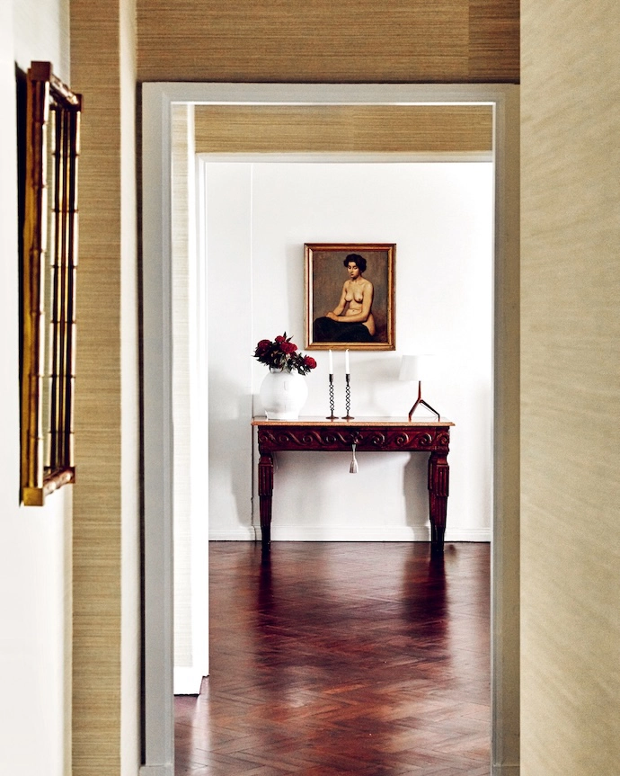 A Phillip Jeffries Manila Hemp grass-cloth wallcovering in Pear lines the corridor to the dining room, where an antique console displays a Foscarini Lumiere lamp beneath a painting that’s a family heirloom.