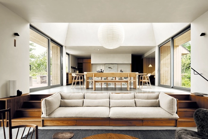 Following the island’s gradual slope, the open-plan kitchen and dining areas step down into a sunken living room. A custom-designed walnut sofa and stairs (the wood chosen to weather with time) bridge the spaces. A bespoke dining table and benches, with Naoto Fukasawa’s Hiroshima armchairs at either end, sit below an Akari pendant.