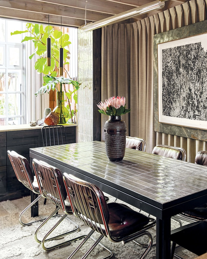 The dining table, built by Jonty from a steel frame and featuring the same tiles used on the walls, anchors the space between the kitchen and the island. Chrome vintage chairs from Cosmic Modern and a textured rug from Haas introduce lighter notes against the weight of the stone.